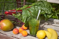 Grüner Smoothie im Glas, umgeben von Mangold, Mango, Aprikosen, Banane und Zitrone auf Holztisch