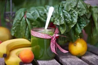 Grüner Smoothie im Glas mit Schleifchen, umgeben von Mangold, Mango, Aprikosen, Banane und Zitrone auf Holztisch